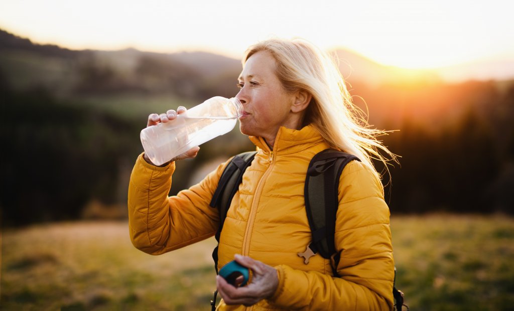 Attractive Senior Woman Walking Outdoors In Nature At Sunset, Drinking Water.