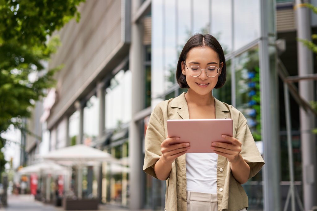 Outdoor Shot Of Young Korean Woman Stands On Street With Laptop, Wears Glasses, Reads, Smiles Happily